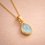 Gold necklace with a teardrop-shaped light blue natural aquamarine gemstone pendant on a beige background