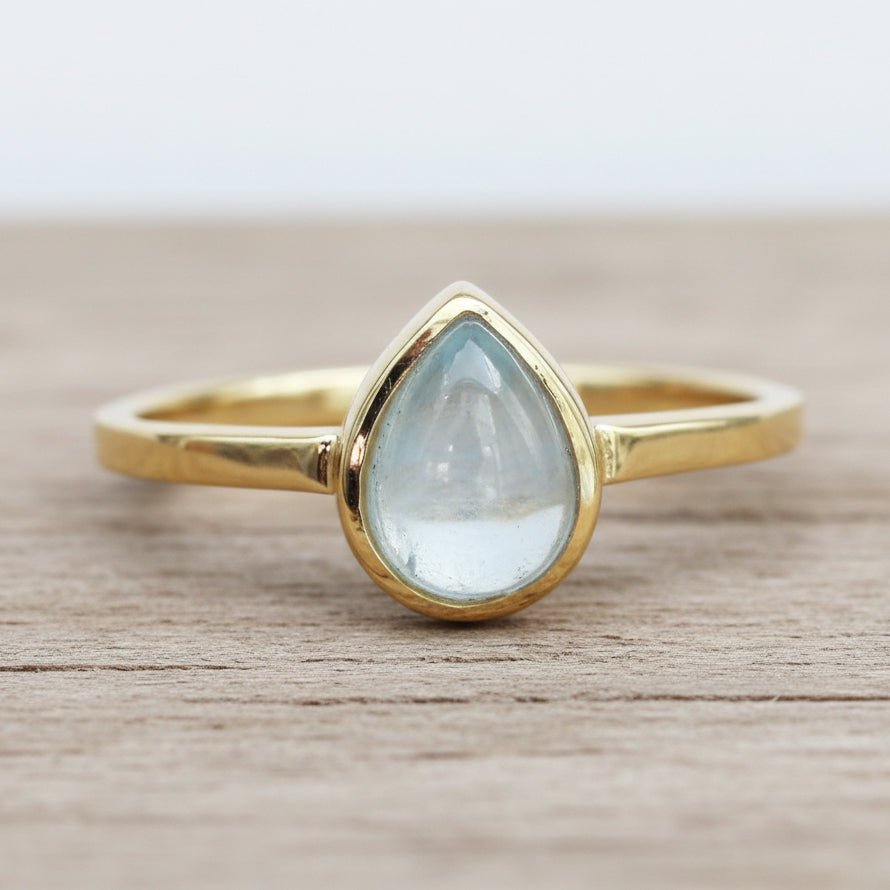 Gold ring with a teardrop-shaped light blue aquamarine gemstone on a wooden surface