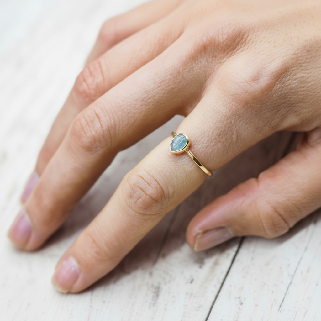 Gold ring with a tear drop shaped aquamarine gemstone on a finger against a light background
