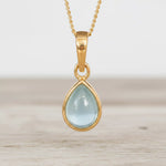 Gold March Birthstone Necklace with a light blue teardrop-shaped natural aquamarine gemstone on a light surface