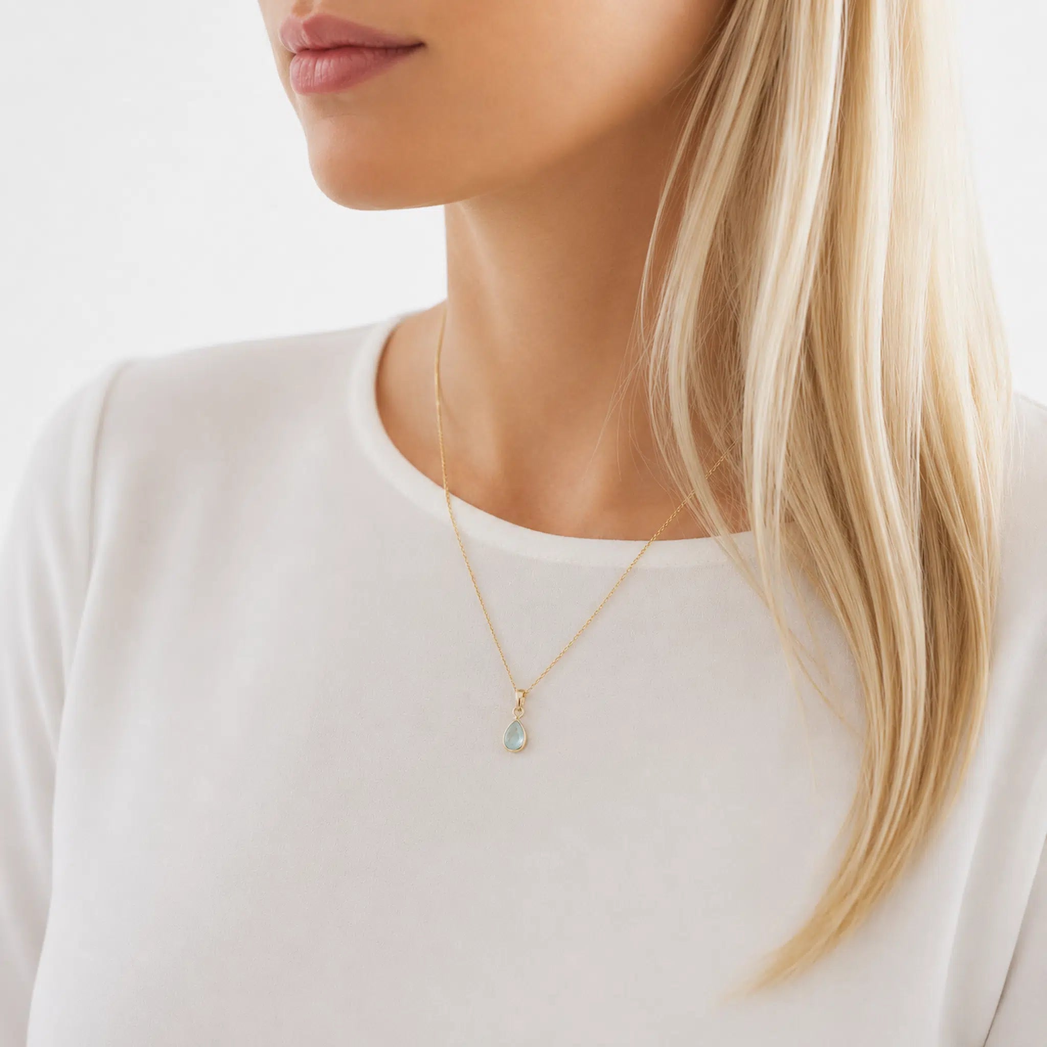 Close-up of a person wearing a gold march birthstone necklace with a pear-shaped natural aquamarine gemstone pendant on a white background