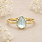 Gold march birthstone ring with a teardrop-shaped light blue aquamarine gemstone on a beige background with delicate flowers.
