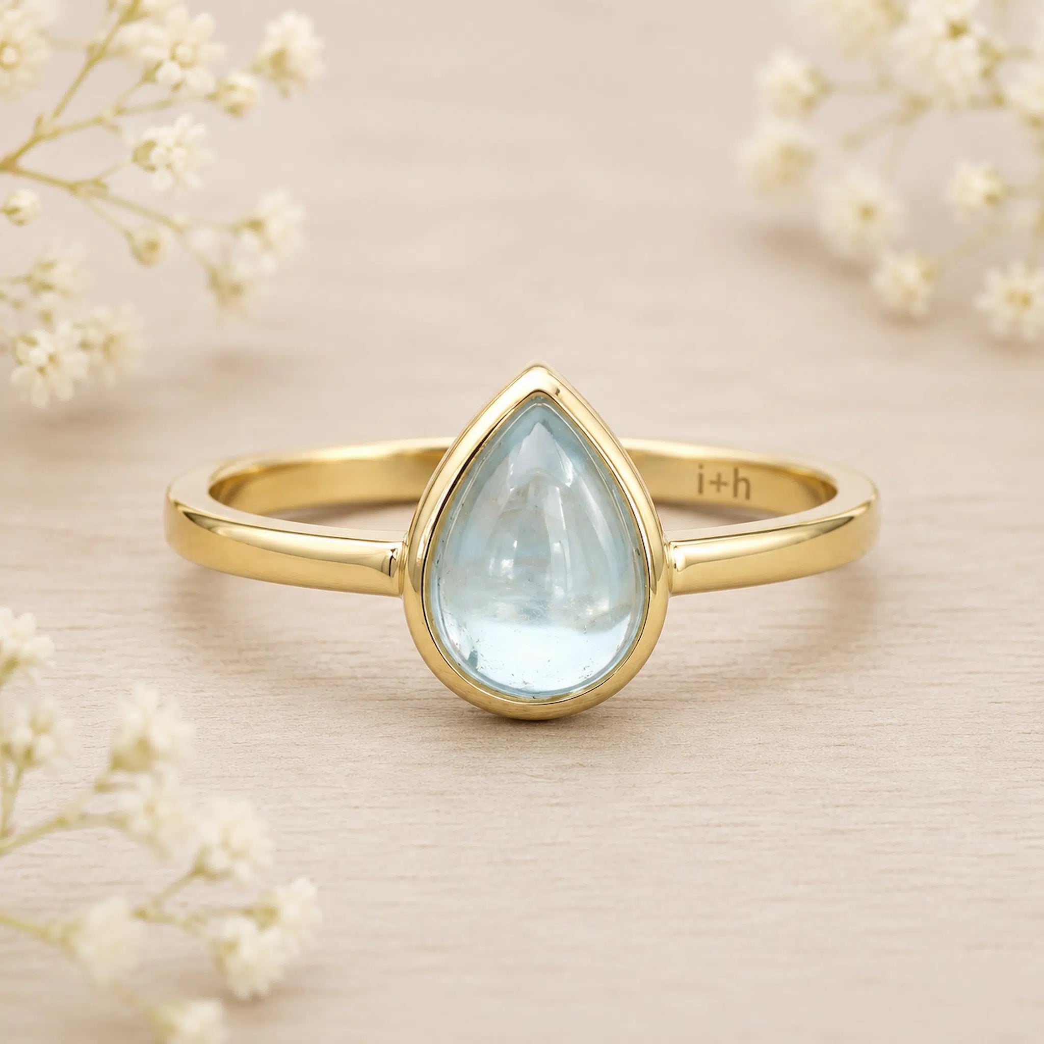 Gold march birthstone ring with a teardrop-shaped light blue aquamarine gemstone on a beige background with delicate flowers.