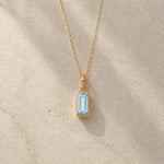 Gold fine chain necklace with a rectangle faceted bezel set march cubic zirconia pendant on a light stone background.