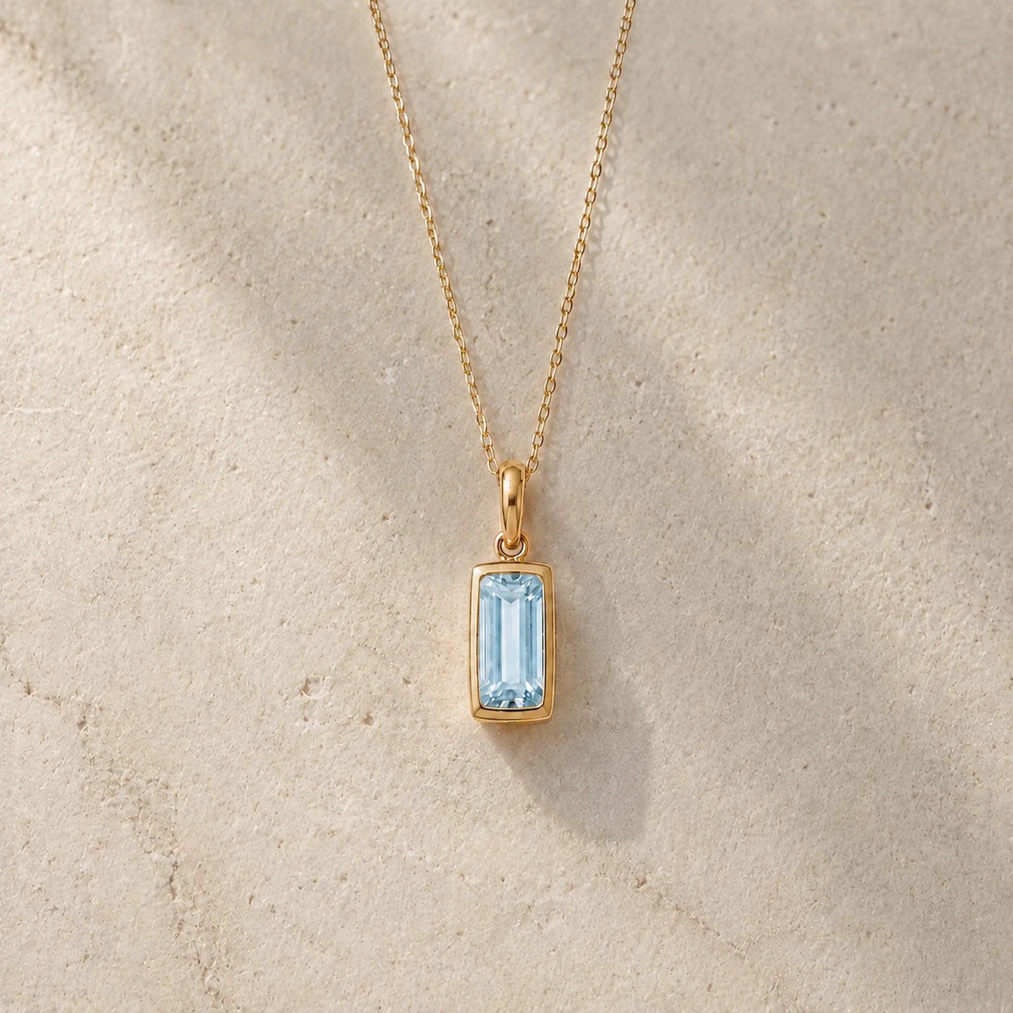 Gold fine chain necklace with a rectangle faceted bezel set march cubic zirconia pendant on a light stone background.
