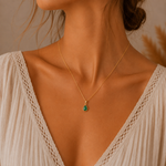 A close-up of a woman wearing a delicate gold chain may birthstone necklace with a small teardrop-shaped natural emerald green pendant, styled with a cream-colored lace top.
