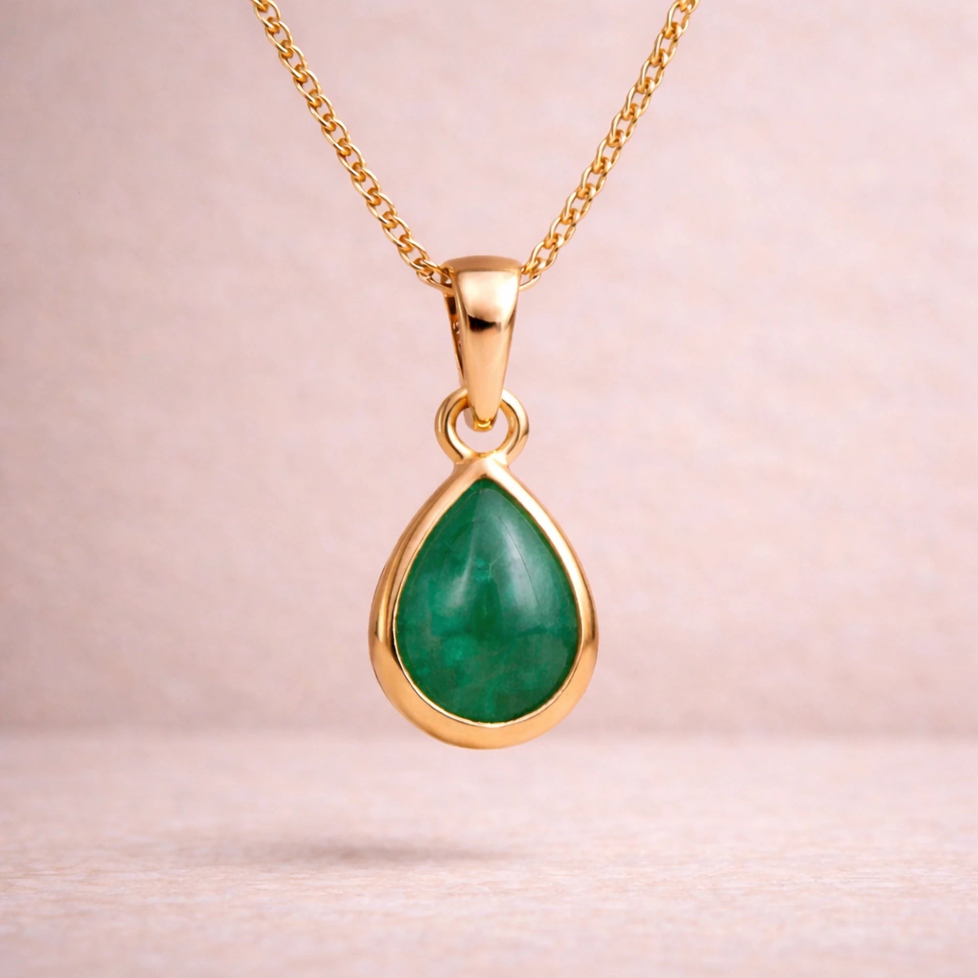 Gold necklace with a green teardrop natural emerald gemstone pendant on a light pink background