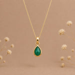 A gold may birthstone necklace with a teardrop pendant necklace with a natural green emerald gemstone, displayed against a warm beige background with small blurred dried flowers.