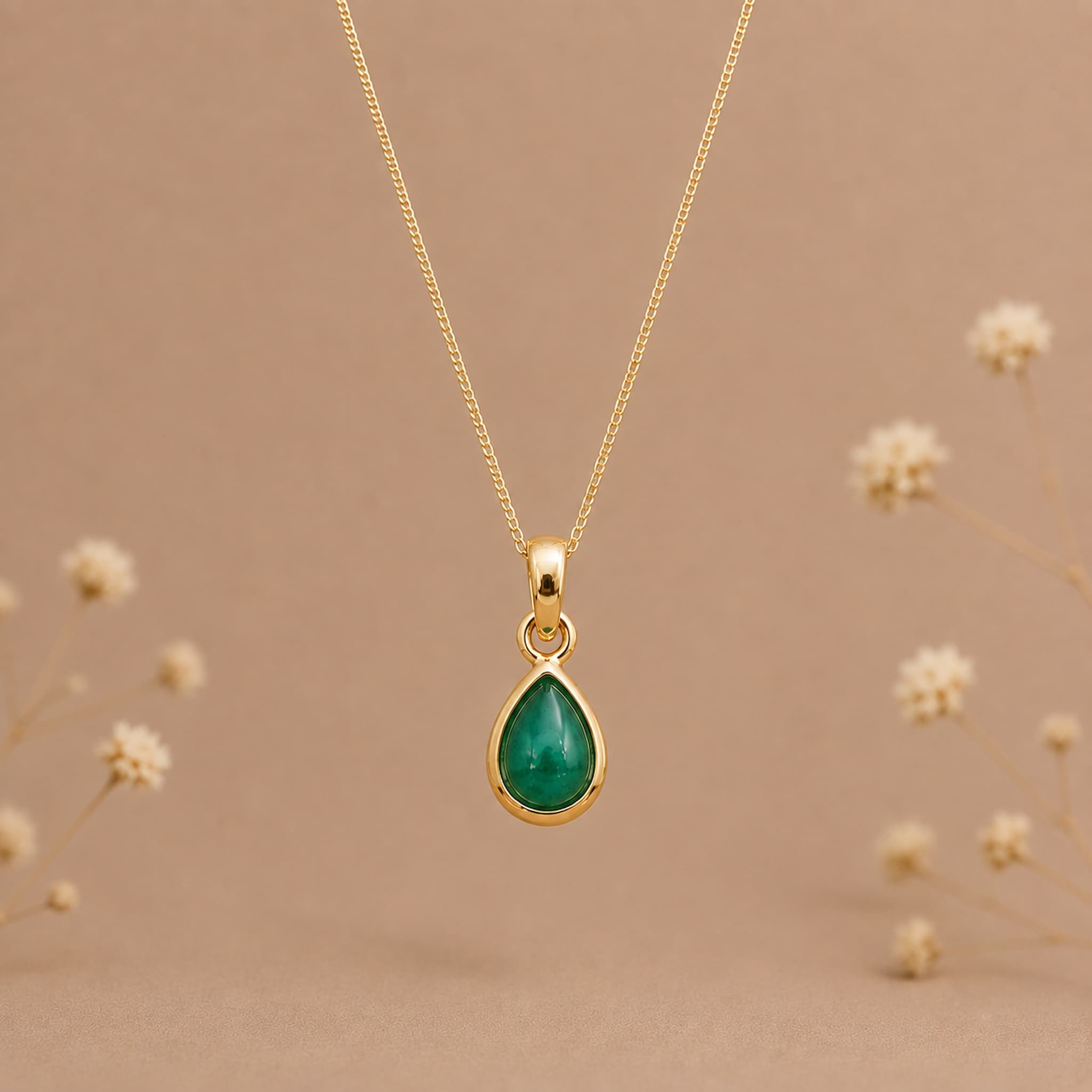 A gold may birthstone necklace with a teardrop pendant necklace with a natural green emerald gemstone, displayed against a warm beige background with small blurred dried flowers.