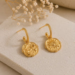 A pair of gold-toned dangle earrings with circular sunburst medallions and textured rope-like hoops, displayed on a light stone slab with dried white flowers in the background.