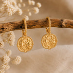 A pair of gold-toned dangle earrings with circular sun-and-moon medallion charms, hanging from a rustic wooden branch with dried white flowers in a soft beige setting.
