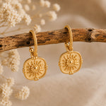 A pair of gold-toned bohemian-style dangle earrings with sunburst medallions, displayed on a rustic wooden branch with dried white flowers against a soft beige fabric background.