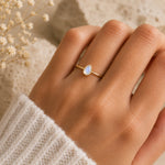 A gold ring with a teardrop-shaped natural moonstone worn on a person's ring finger, shown against a soft beige background with dried flowers and a cream knit sleeve.