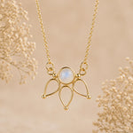 A gold pendant necklace featuring a round iridescent moonstone surrounded by three teardrop-shaped gold loops, hanging against a soft beige background with delicate dried flowers.
