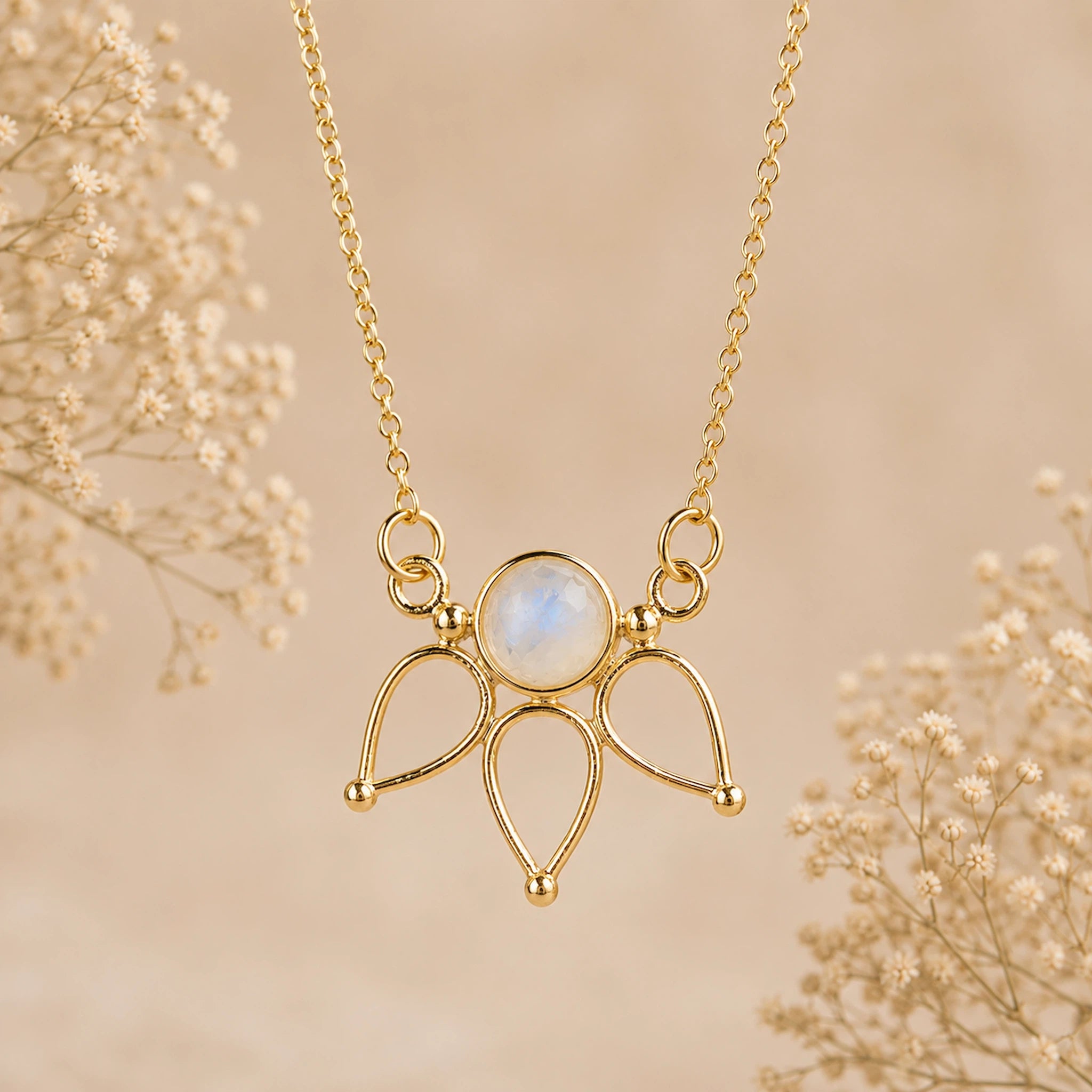 A gold pendant necklace featuring a round iridescent moonstone surrounded by three teardrop-shaped gold loops, hanging against a soft beige background with delicate dried flowers.