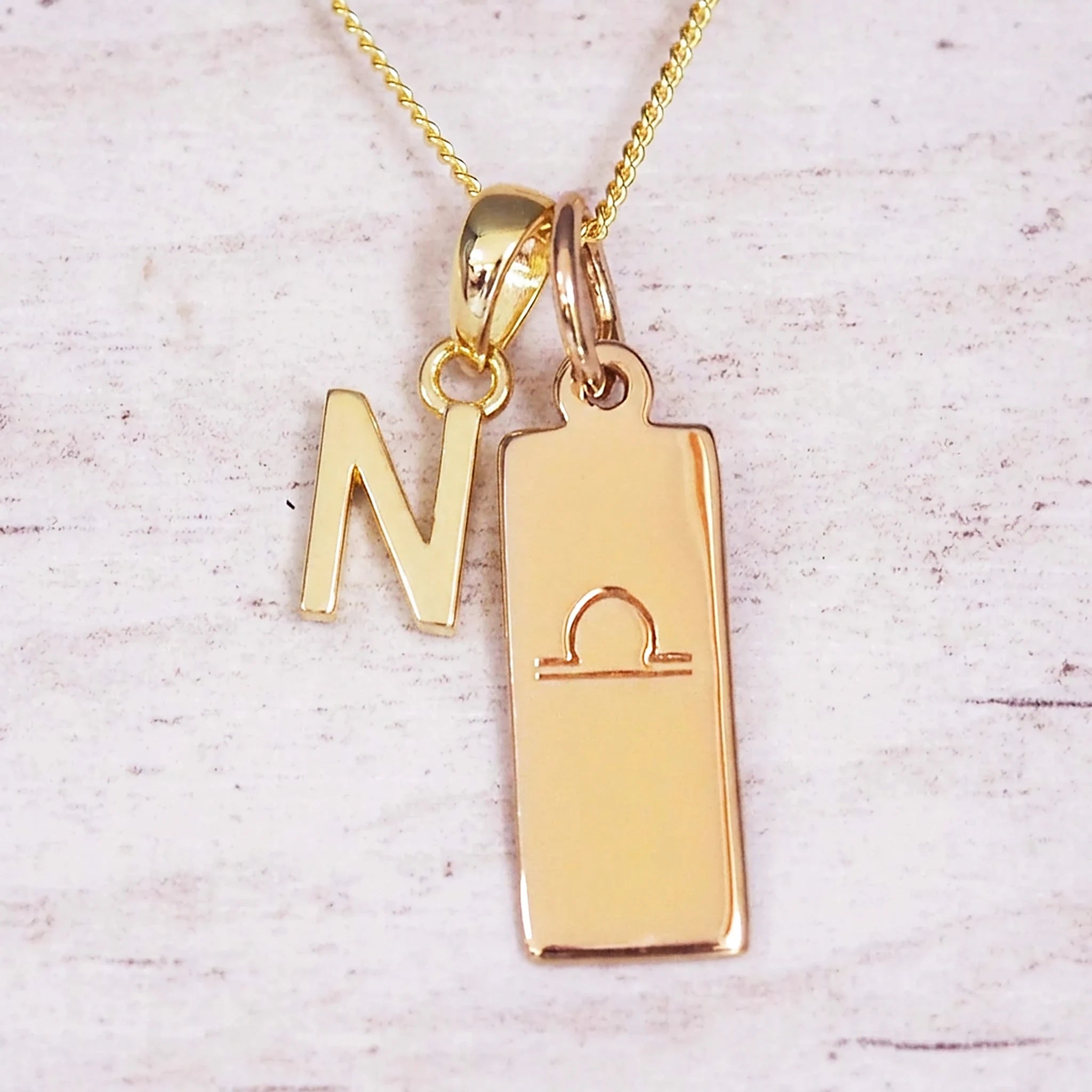 Gold initial and Libra zodiac necklace with a letter N charm and engraved bar pendant on whitewashed wood