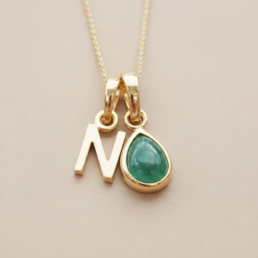 Gold necklace with 'N' pendant and green teardrop charm on a beige background