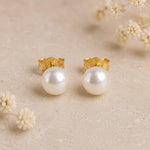 A pair of gold pearl earrings with large round white freshwater pearl studs, displayed on a textured beige surface with small dried flowers in the background.