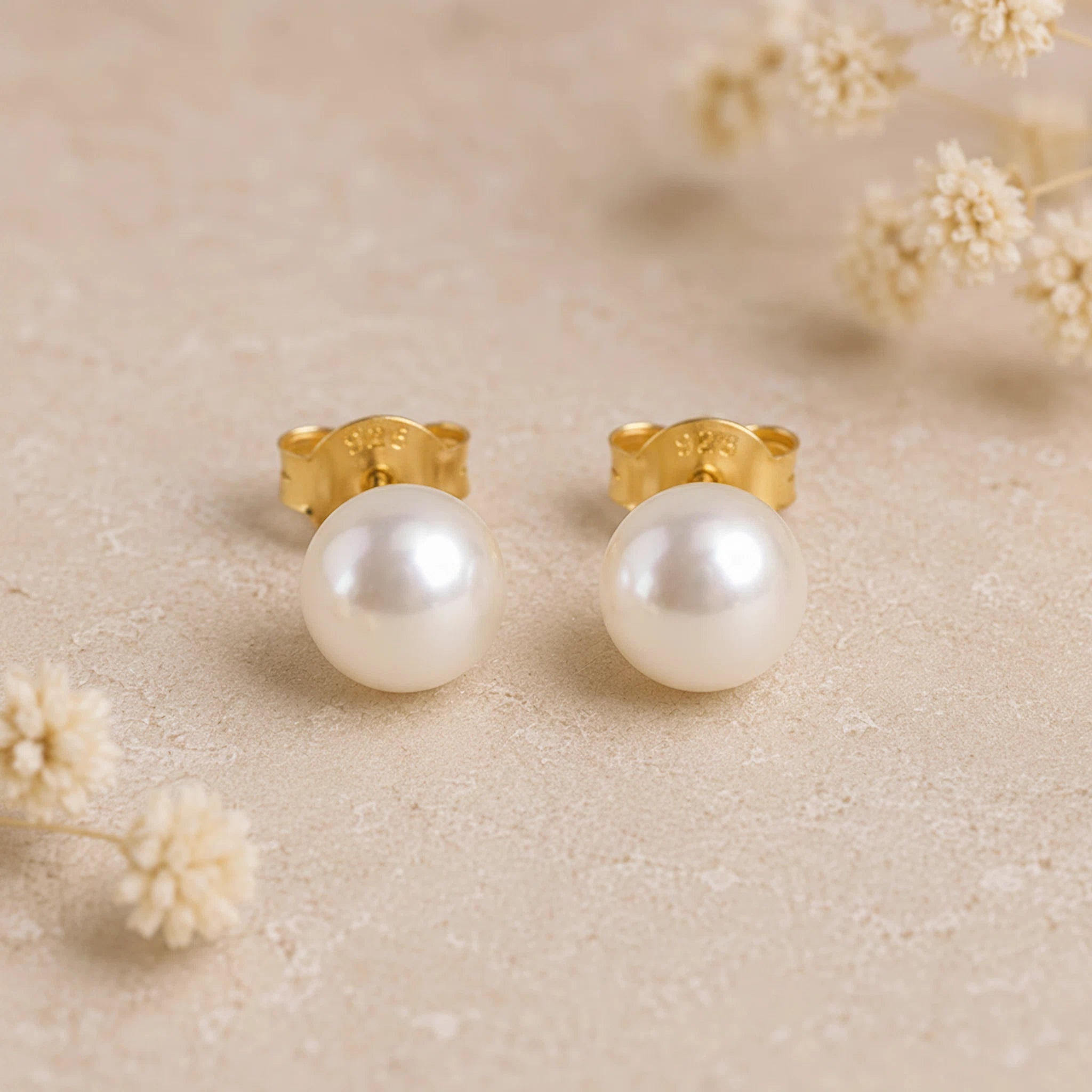 A pair of gold pearl earrings with large round white freshwater pearl studs, displayed on a textured beige surface with small dried flowers in the background.