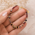 A delicate beaded bracelet with gold-toned chain and charms, held in a hand. The bracelet features small maroon beads, gold spacers, and various colored gemstone beads, including a marquise-cut green stone charm.