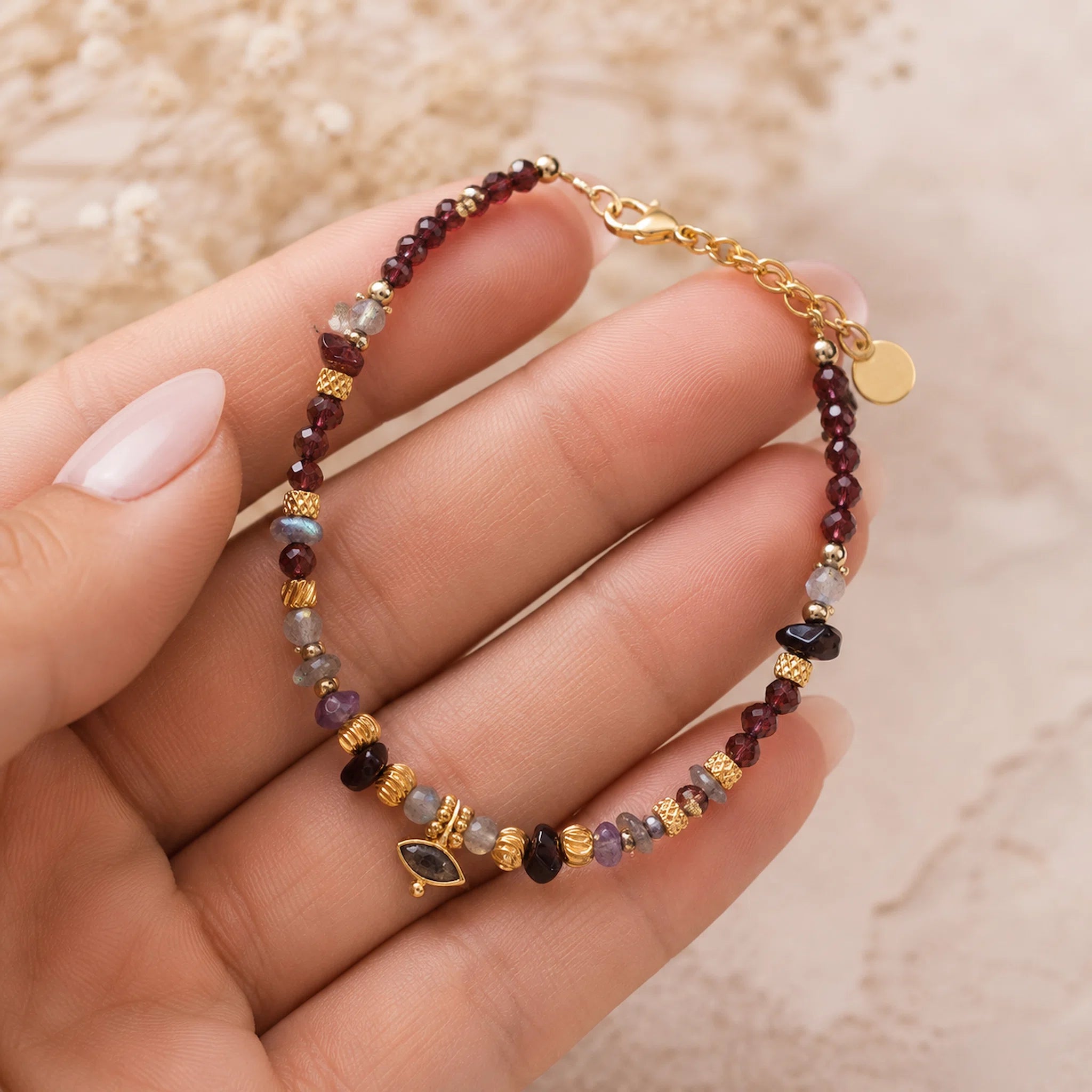 A delicate beaded bracelet with gold-toned chain and charms, held in a hand. The bracelet features small maroon beads, gold spacers, and various colored gemstone beads, including a marquise-cut green stone charm.