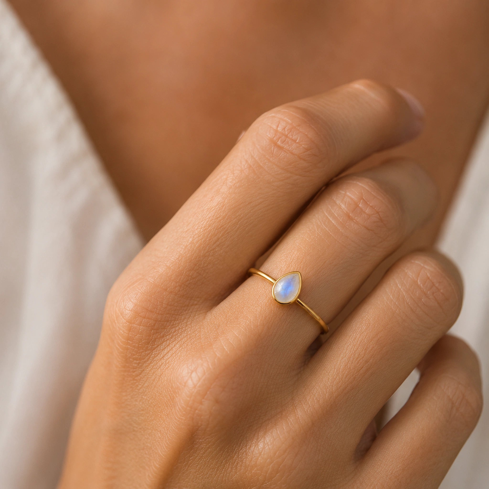 A delicate gold ring with a small teardrop-shaped moonstone set on a finger, shown in a close-up lifestyle shot against a soft white garment and warm skin tones.