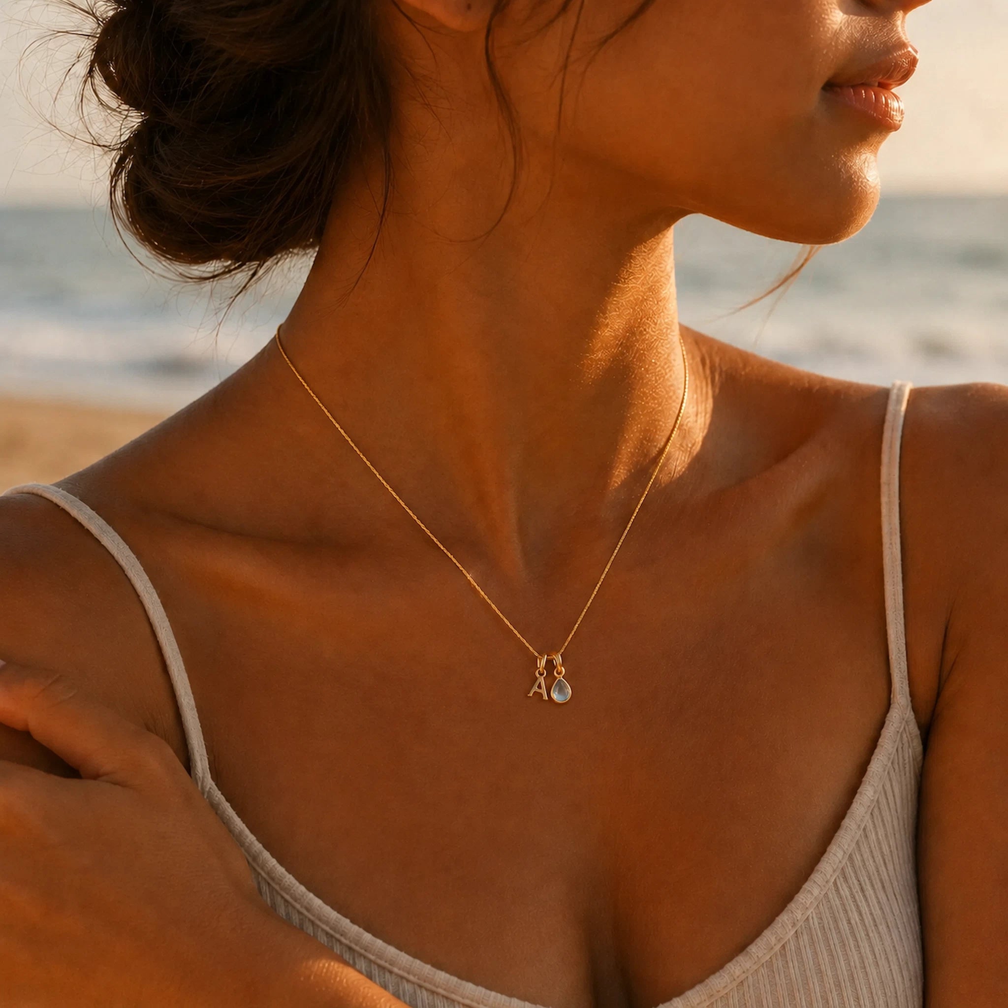 Woman on a beach wearing a gold fine chain necklace with a A letter pendant and a pear shaped november birthstone blue topaz gemstone pendant