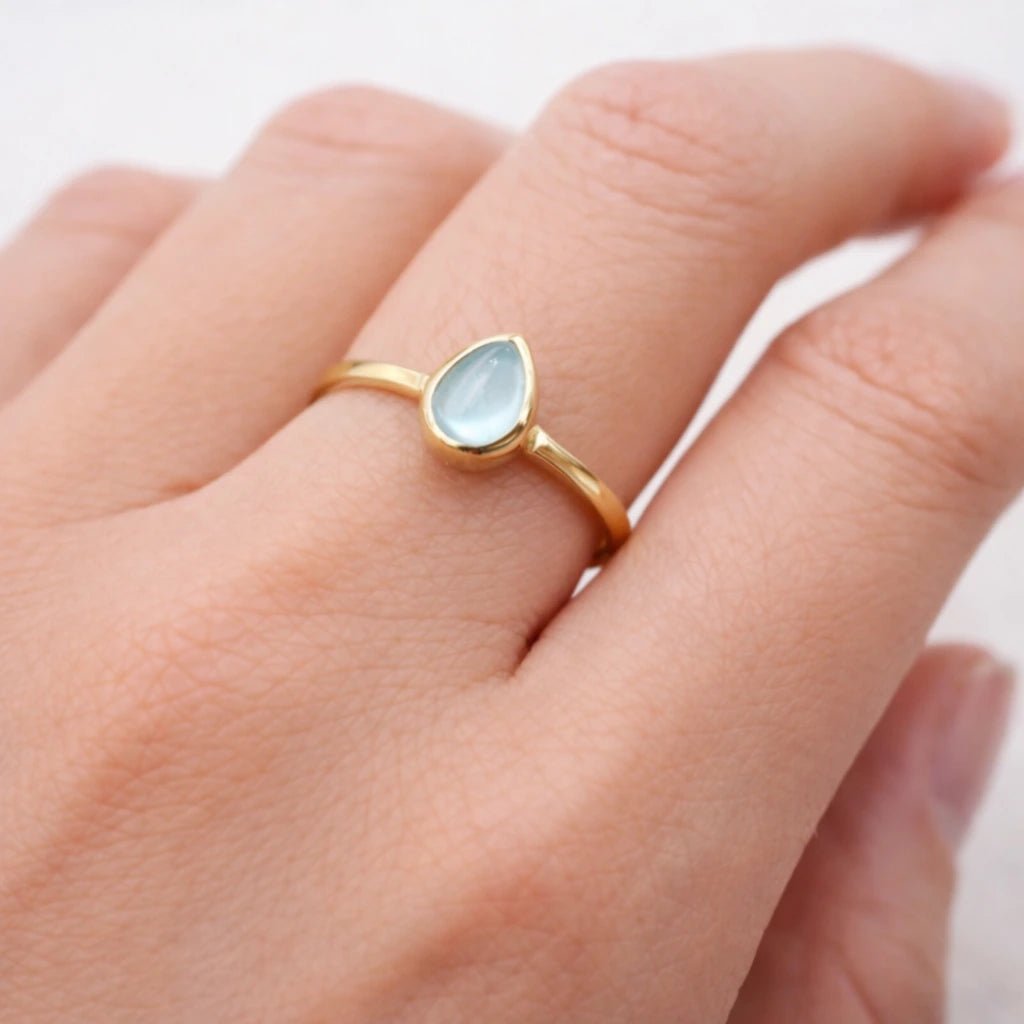 Gold ring with a light blue natural topaz gemstone on a person's finger against a white background