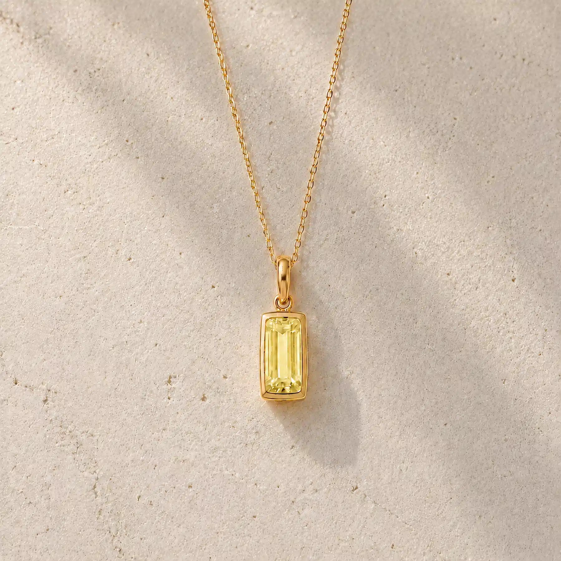 Gold fine chain necklace with a rectangle faceted bezel set november cubic zirconia pendant on a light stone background.