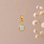 A gold teardrop pendant necklace with an iridescent opal stone, hanging against a soft beige background with blurred dried flowers on the right.