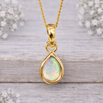 Gold pendant with a teardrop-shaped opal stone on a wooden surface with white flowers.