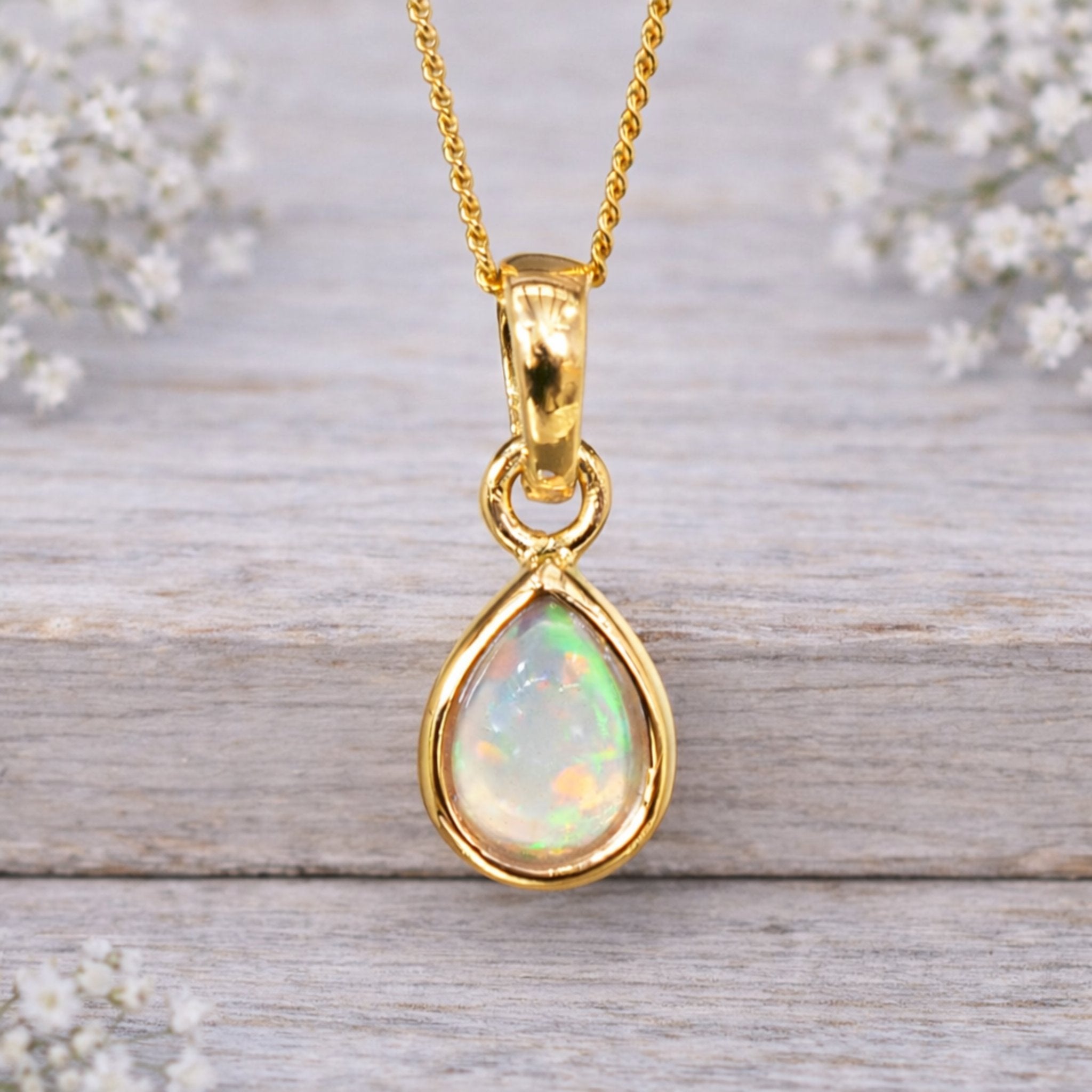 Gold pendant with a teardrop-shaped opal stone on a wooden surface with white flowers.