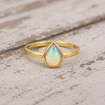 A gold ring with a teardrop-shaped iridescent opal gemstone, resting on a rustic, distressed white wooden surface.