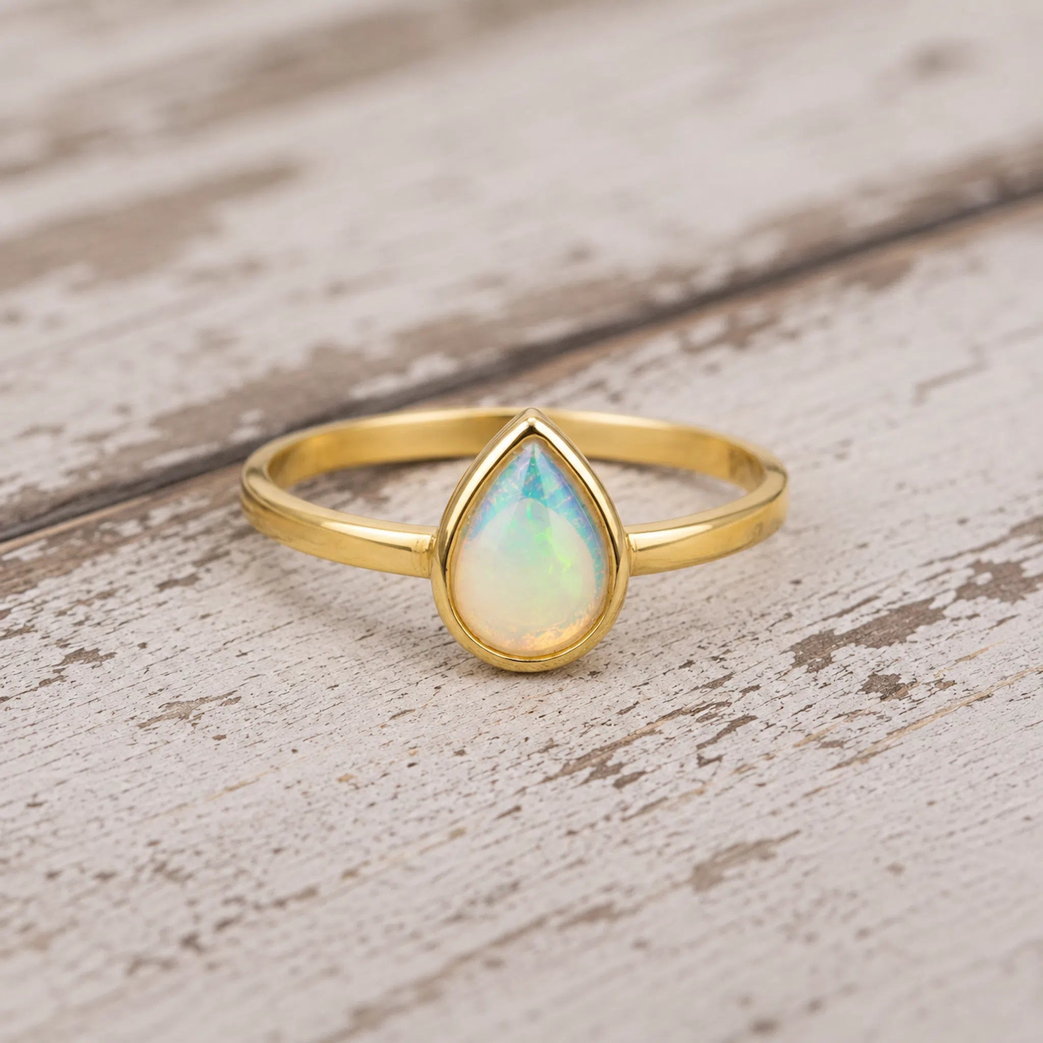A gold ring with a teardrop-shaped iridescent opal gemstone, resting on a rustic, distressed white wooden surface.
