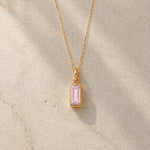 Gold fine chain necklace with a rectangle faceted bezel set october cubic zirconia pendant on a light stone background.