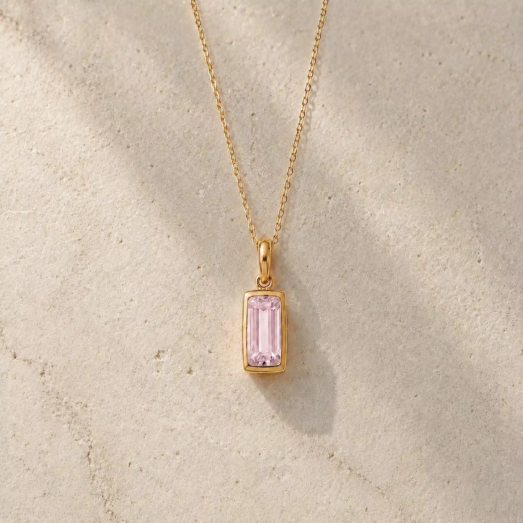 Gold fine chain necklace with a rectangle faceted bezel set october cubic zirconia pendant on a light stone background.