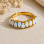 A gold ring featuring a row of teardrop-shaped white opal gemstones, displayed on a textured beige surface with dried flowers and a soft beige fabric in the background.
