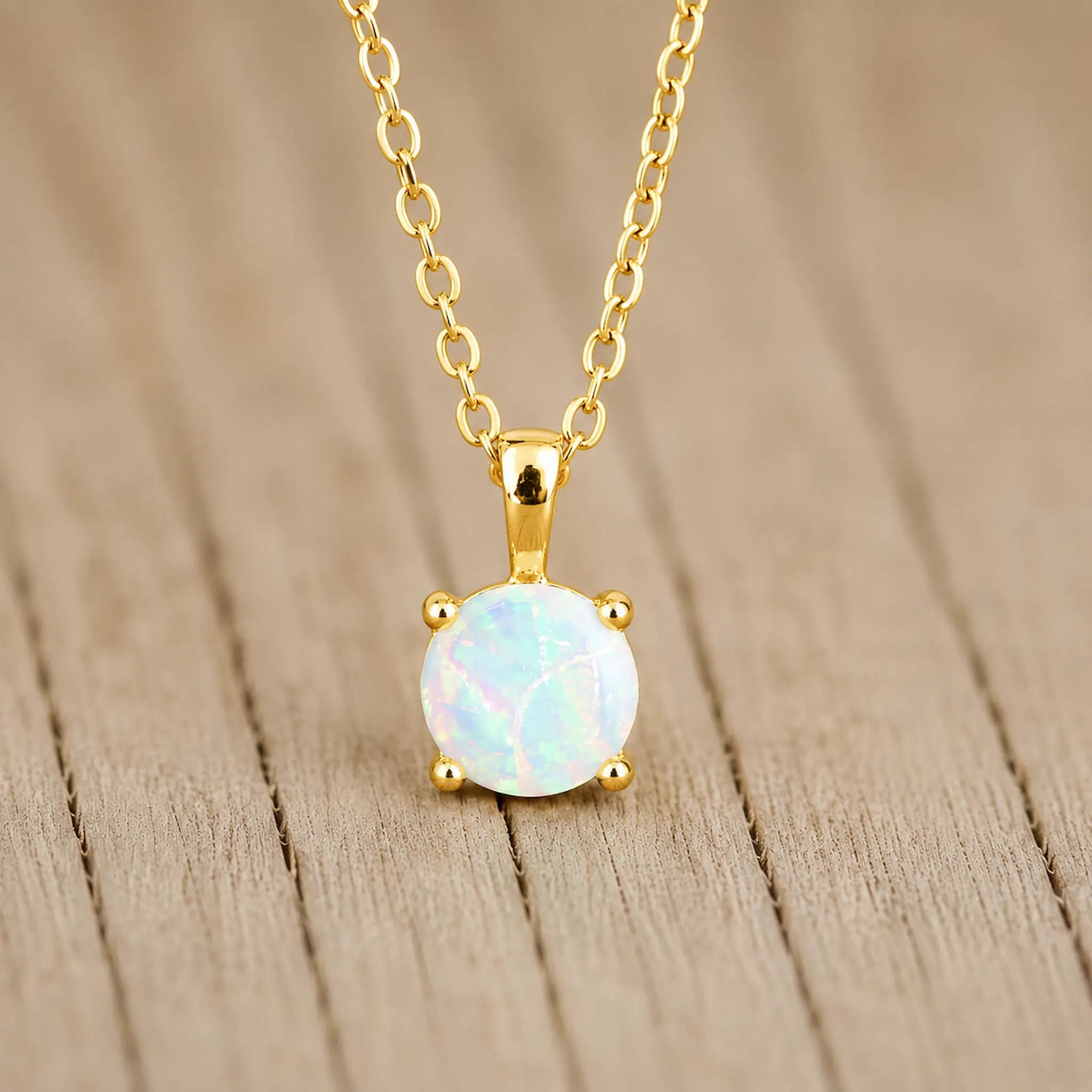 A gold pendant necklace featuring a round white opal stone in a four-prong setting, hanging from a gold chain against a light brown wooden background.