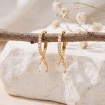 Gold opal teardrop huggie earrings hanging from a rustic branch over white stone