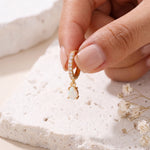 Gold opal teardrop huggie earrings, one held in fingertips over white stone