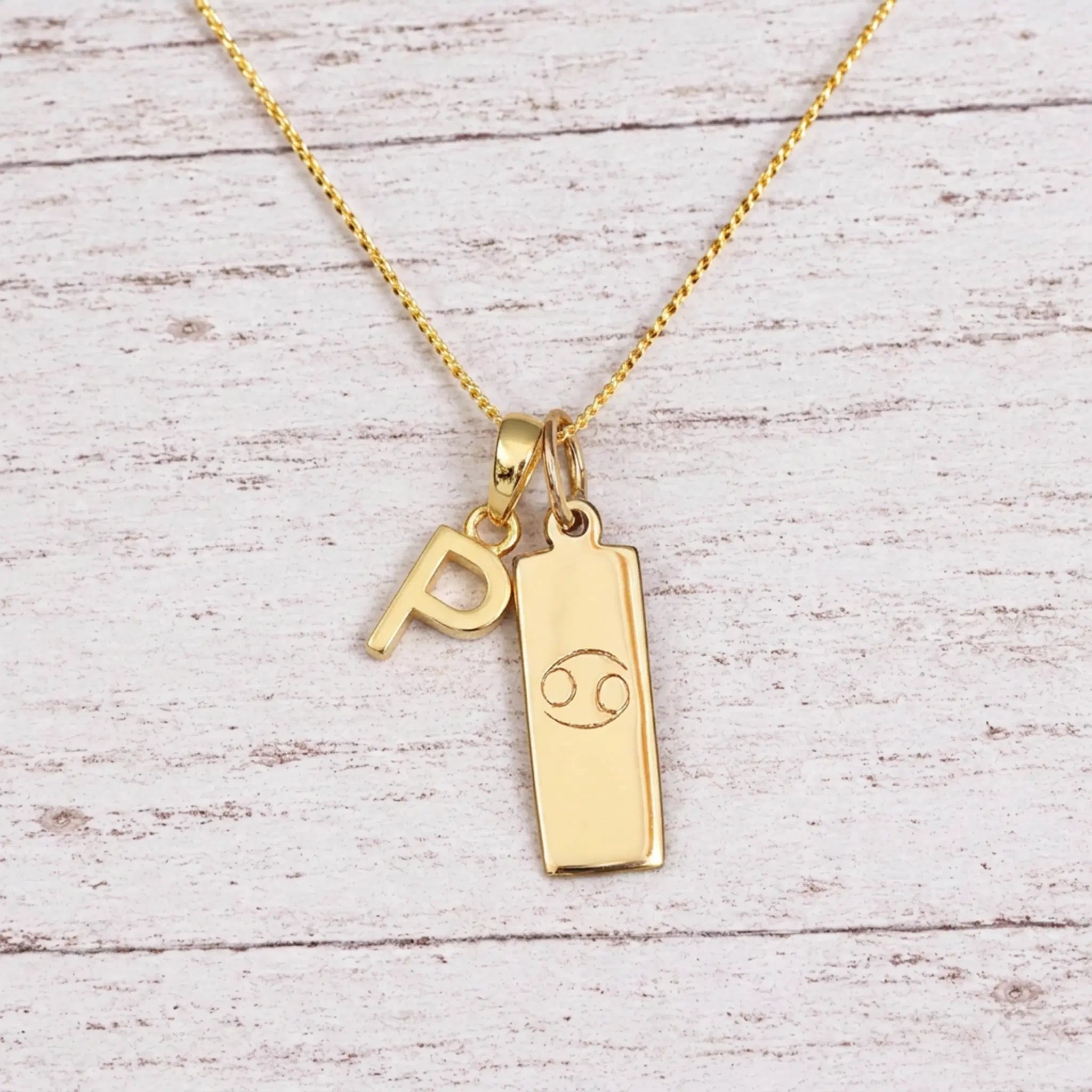 Gold Initial P Pendant and Cancer Zodiac Pendant Necklace on a fine gold chain against a light wooden background