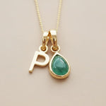 Gold necklace with 'P' and green teardrop pendant on a beige background