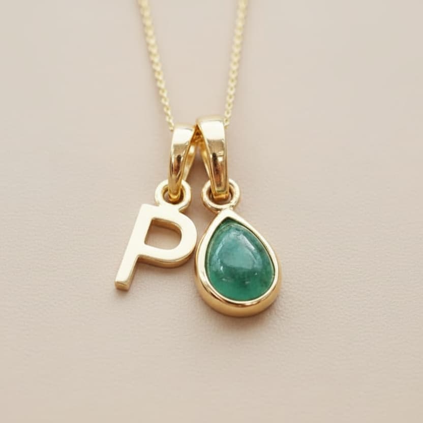 Gold necklace with 'P' and green teardrop pendant on a beige background