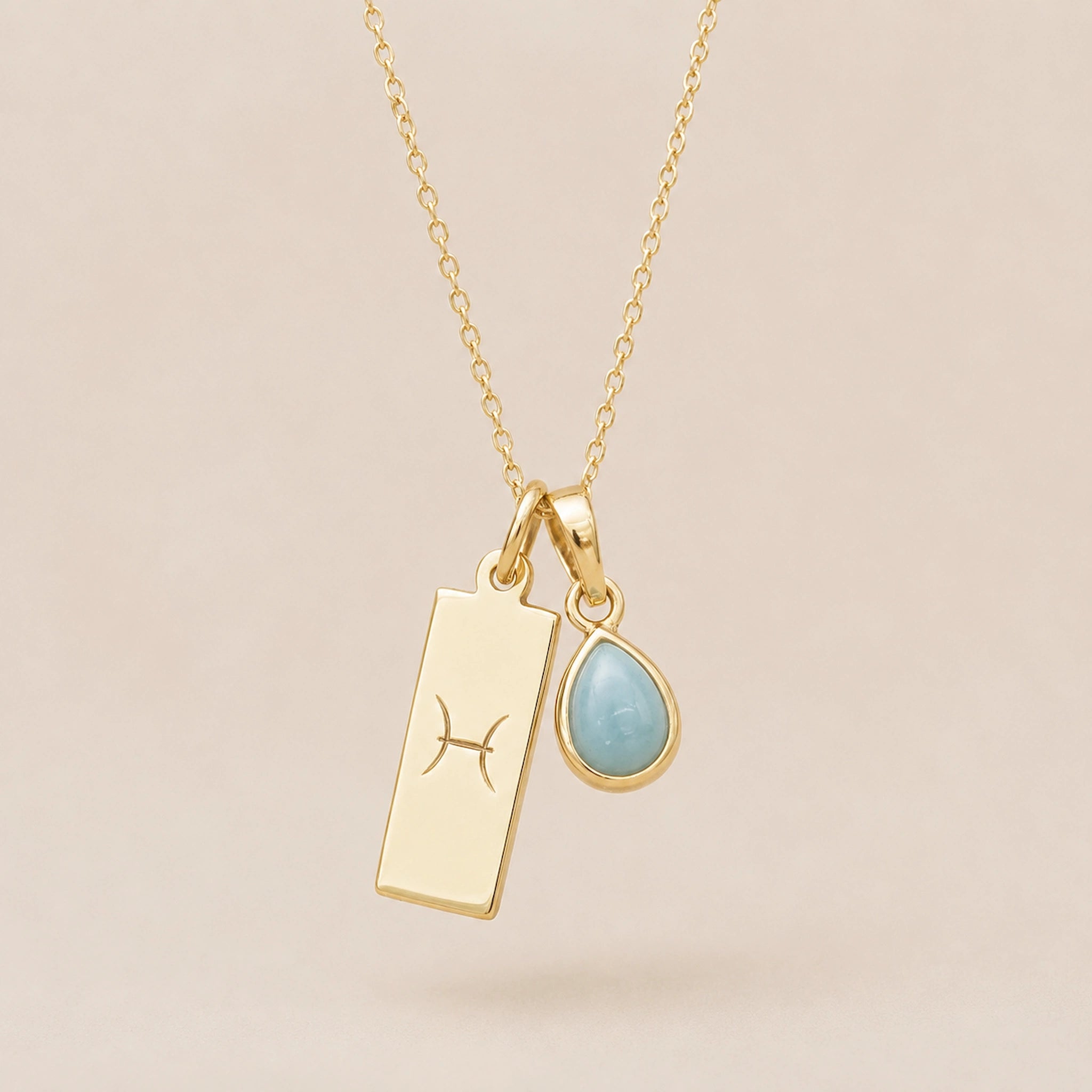 Gold Necklace with teardrop March Birthstone Natural Aquamarine pendant and a rectangle pendant wth Pisces line glyph hanging on a light background.