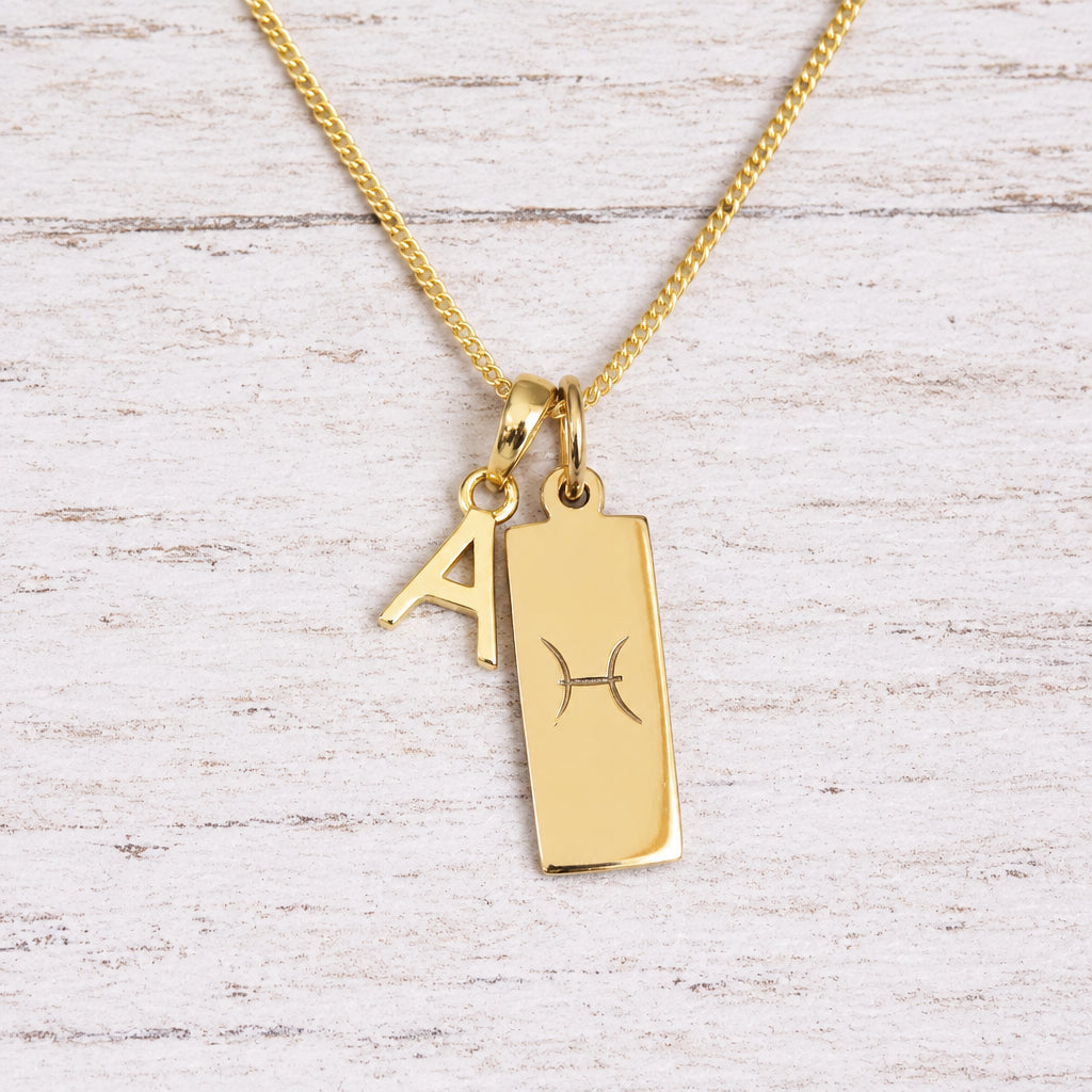 Gold Pisces initial necklace with a letter A charm and engraved zodiac bar on white wood, close-up