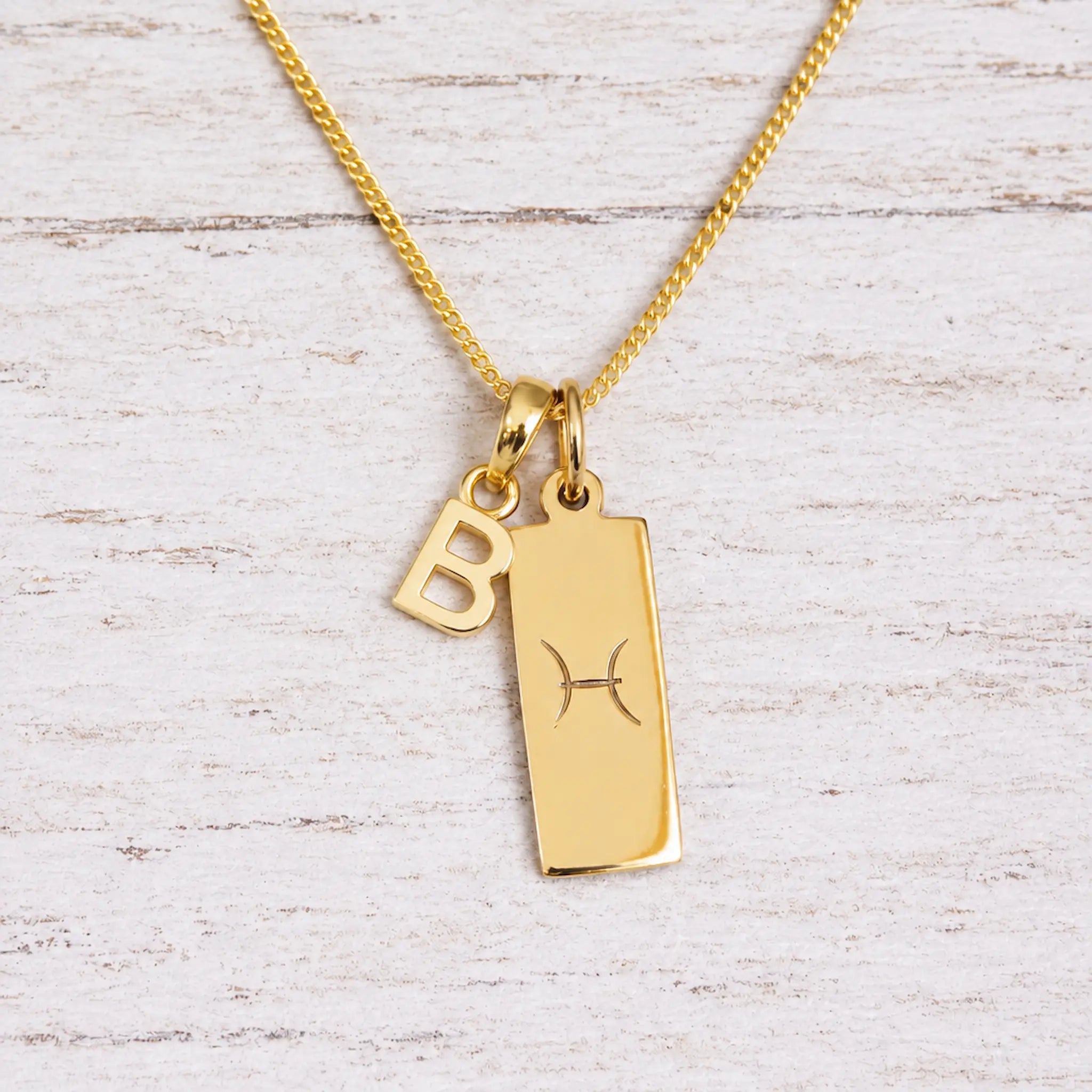 Gold Pisces initial necklace with a letter B charm and engraved zodiac bar on white wood, close-up