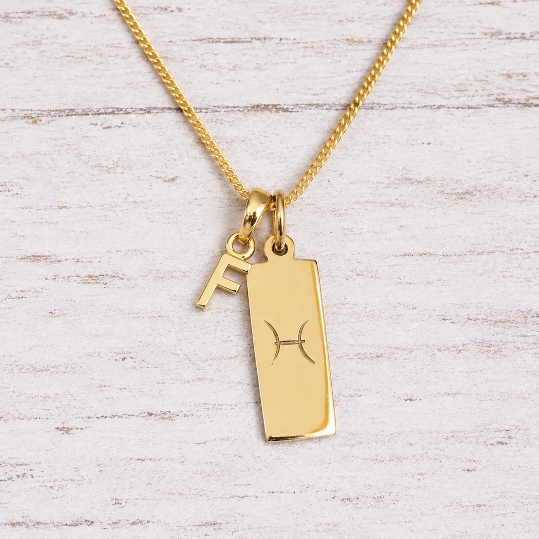 Gold Pisces initial necklace with a letter F charm and engraved zodiac bar on white wood, close-up