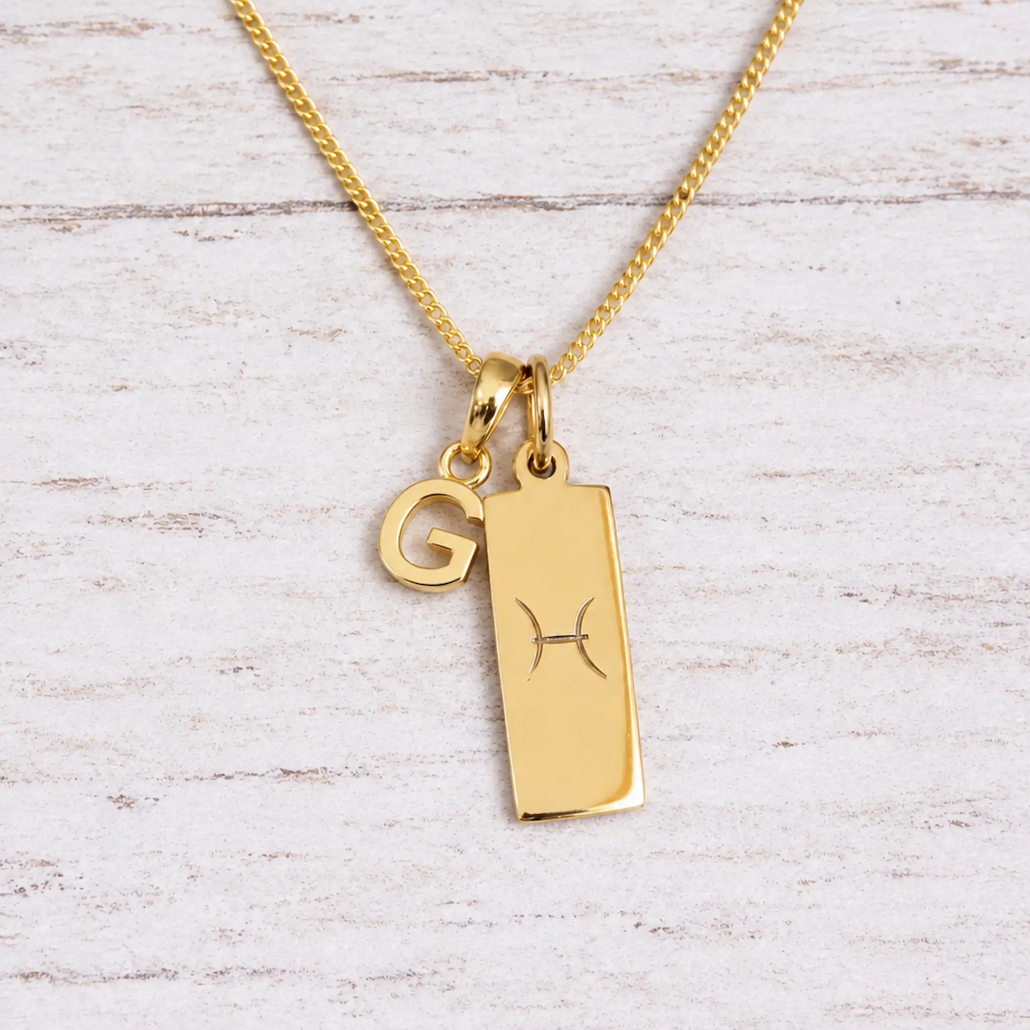 Gold Pisces initial necklace with a letter G charm and engraved zodiac bar on white wood, close-up