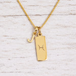 Gold Pisces initial necklace with a letter J charm and engraved zodiac bar on white wood, close-up