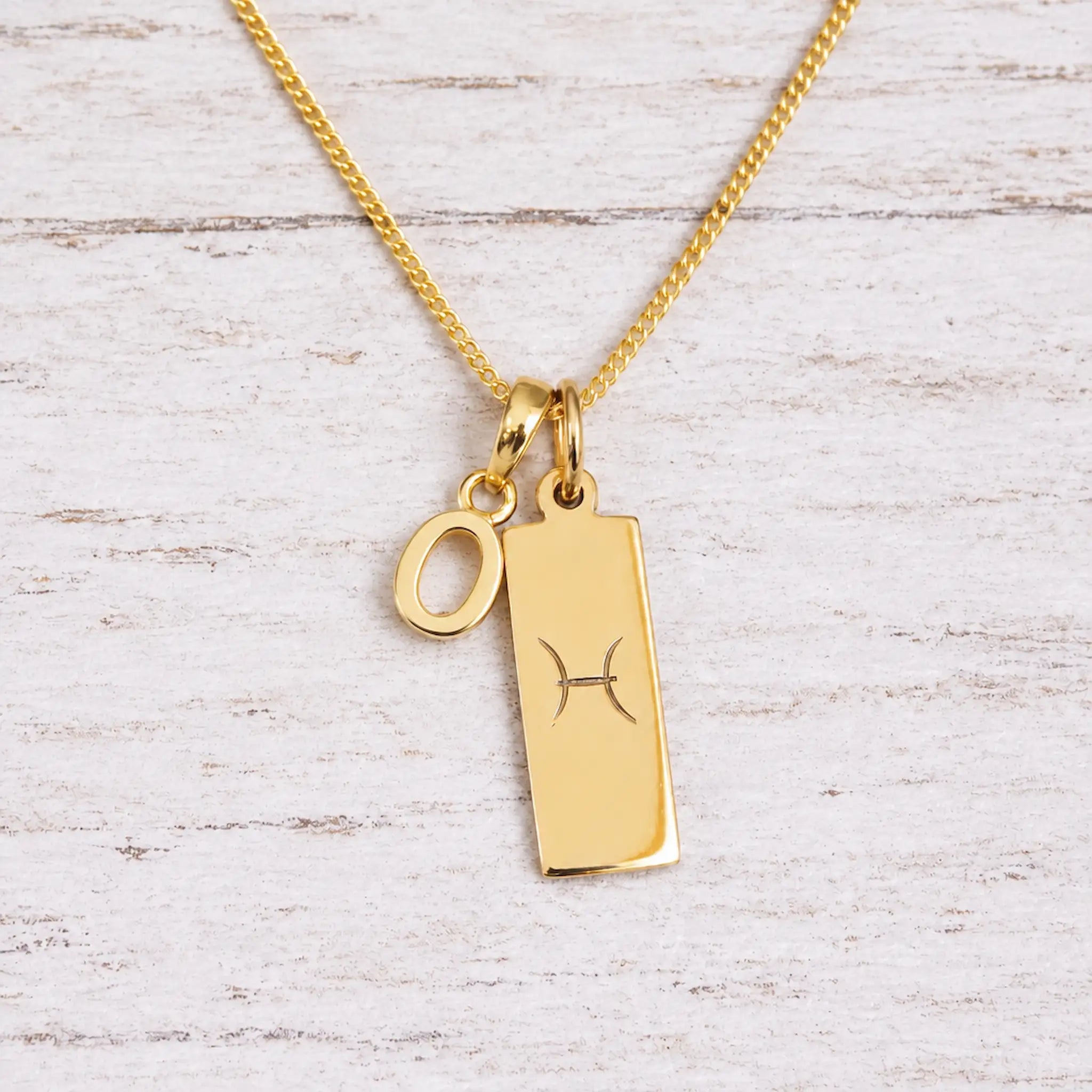 Gold Pisces initial necklace with a letter O charm and engraved zodiac bar on white wood, close-up